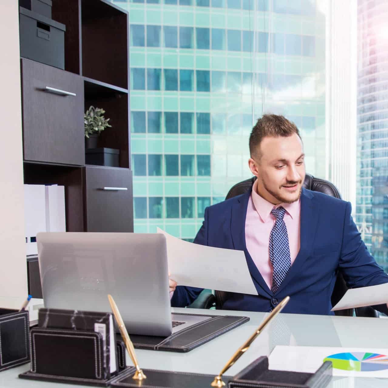 Portrait of smiling handsome businessman working with documents, laptop at the desk in modern office. Marketing manager