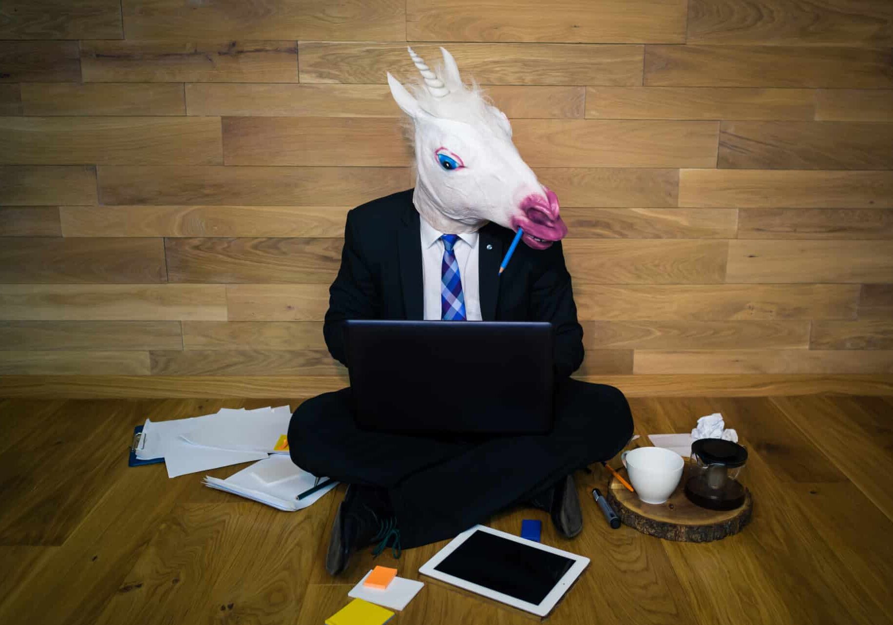 Young man in funny horse mask sits on the floor near a wall and works with laptop. Serious unicorn in a suit and tie enthusiastically works at home office with a pencil in the teeth Marketing Unicorn